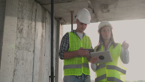 Engineers-designers-a-man-and-a-woman-standing-on-the-roof-of-a-building-under-construction-and-discuss-the-plan-and-progress-of-construction-using-a-tablet-and-mobile-phone.-Modern-builders-discuss-the-infrastructure-of-the-building-and-the-surrounding-area